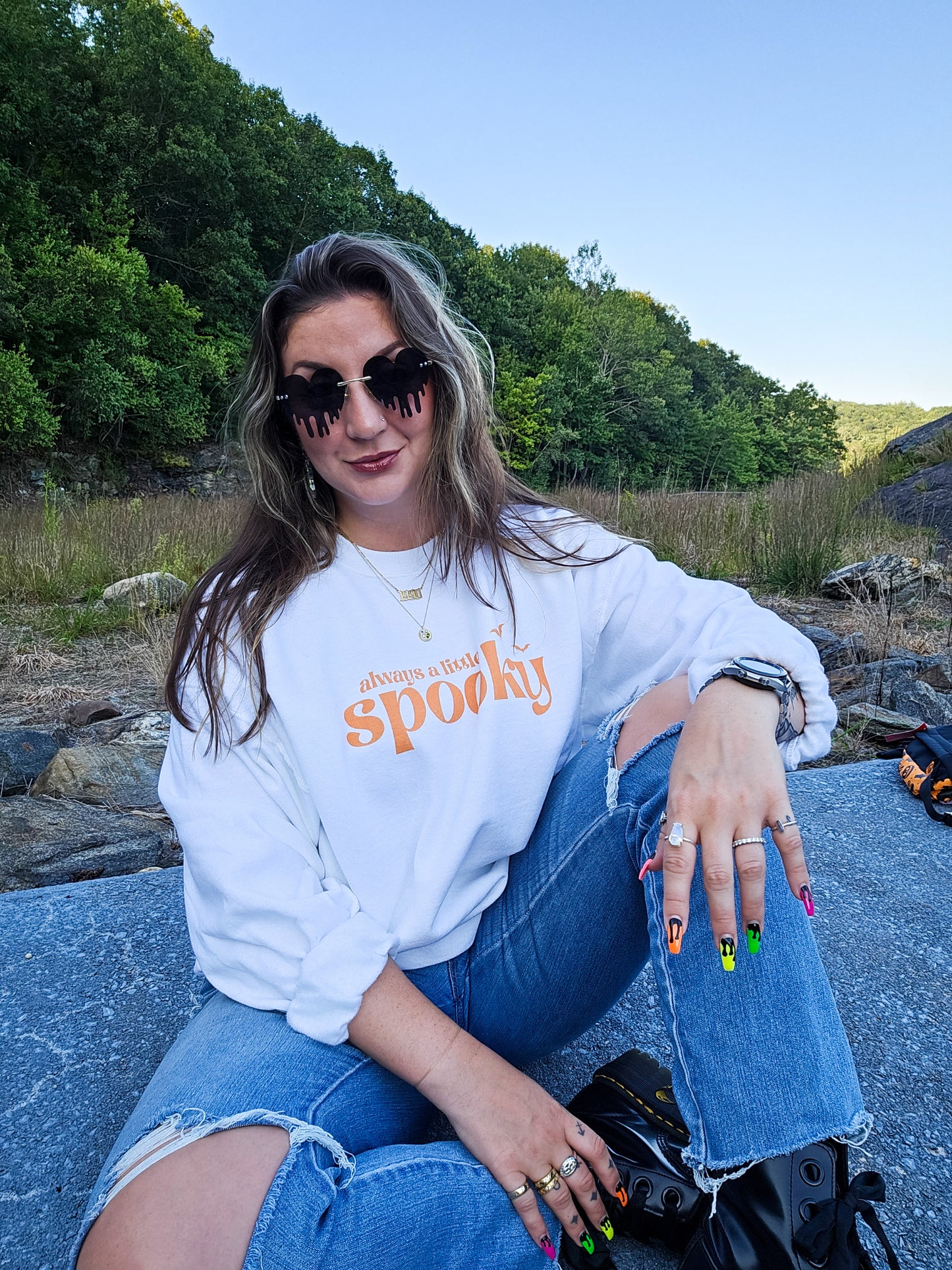 A girl wearing ripped jeans, Dr. Marten boots, black sunglasses, and a white Halloween crewneck sweatshirt that says "always a little spooky" in a trendy orange font