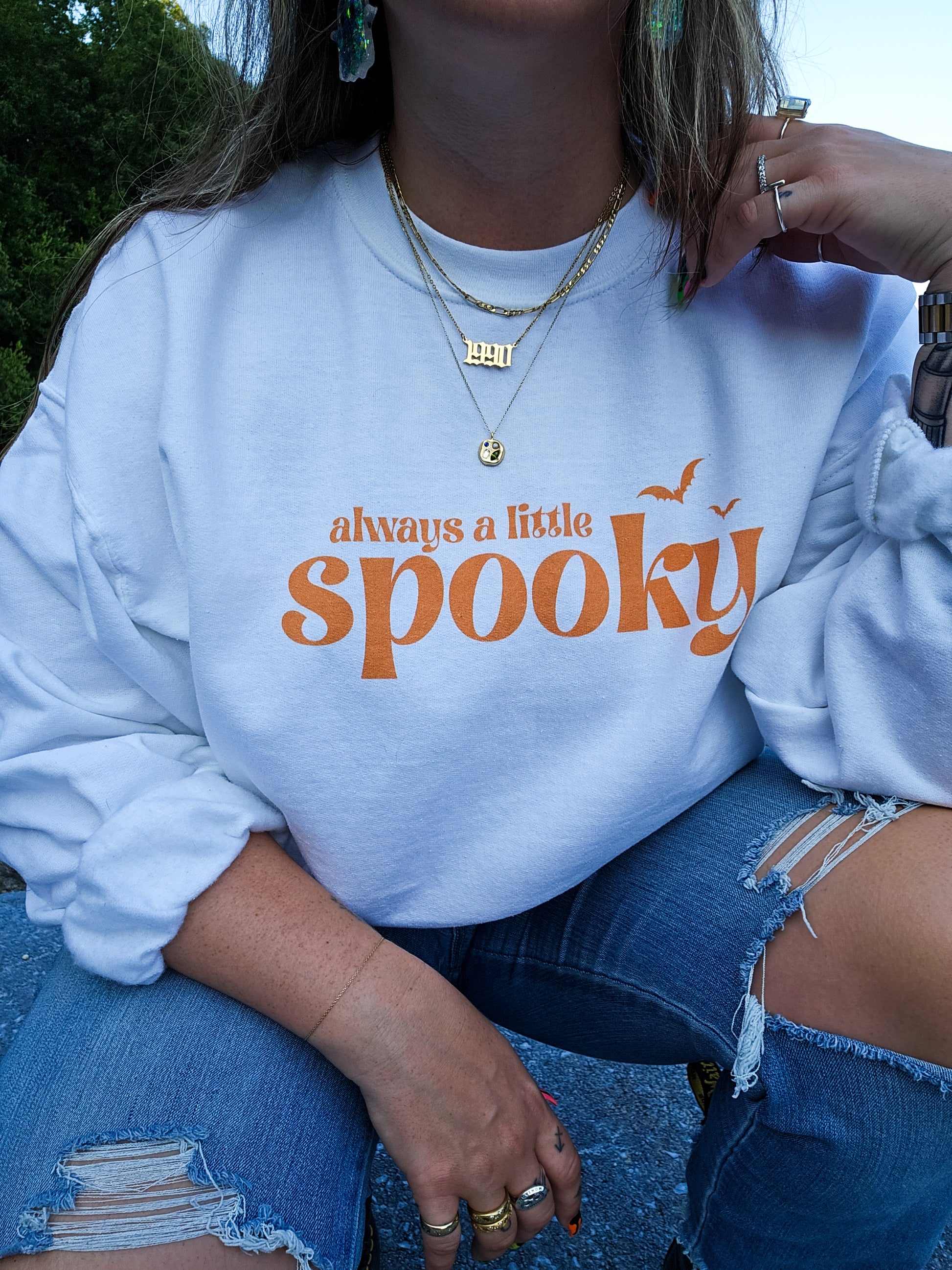 A girl wearing a white Halloween crewneck sweatshirt featuring the phrase "always a little spooky" in a trendy orange font
