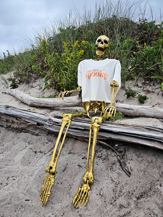 A gold, life-sized skeleton, sitting on a large piece of driftwood, wearing a cropped t-shirt that says "always a little spooky" in a trendy orange font