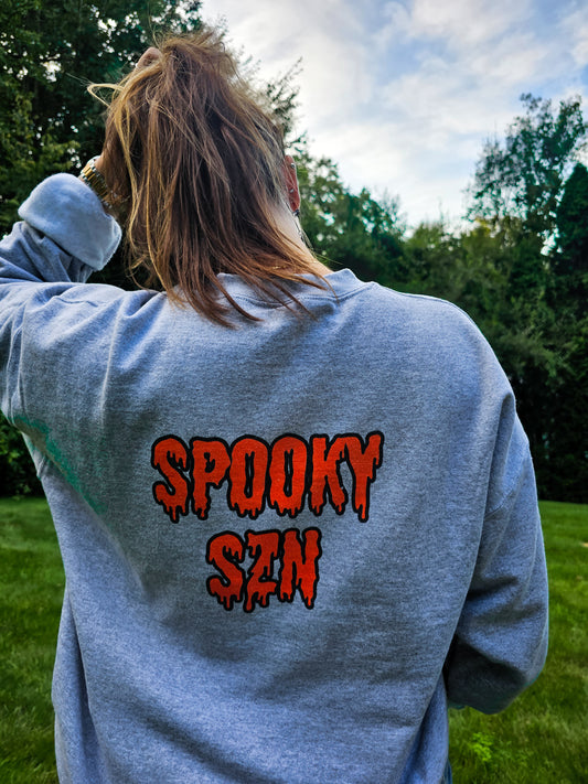 A girl wearing a gray crewneck sweatshirt that says "spooky szn" on the back in a creepy, dripping orange font