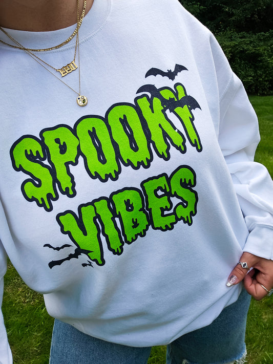 A girl wearing a white Halloween crewneck sweatshirt that says "spooky vibes" in a creepy, dripping lime green font, surrounded by bats