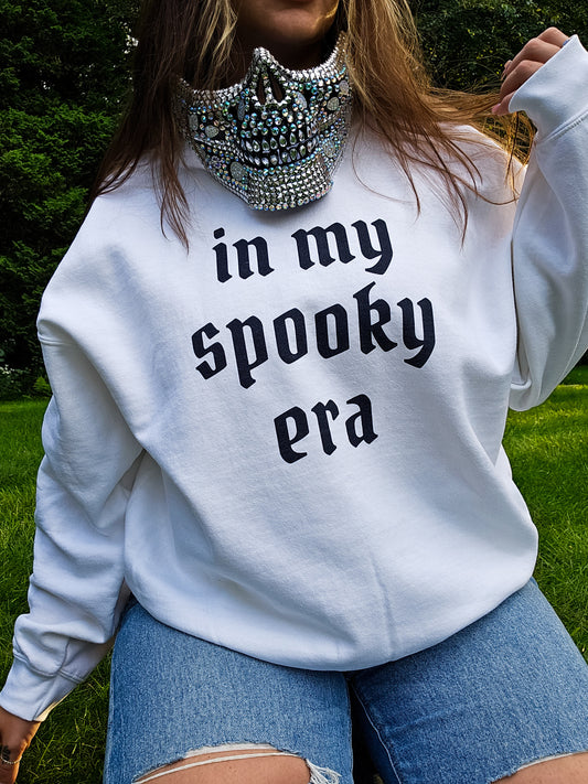 A girl kneeling in the grass, wearing ripped jeans, a bedazzled half-skull mask, and a white sweatshirt that says "in my spooky era" in black gothic font