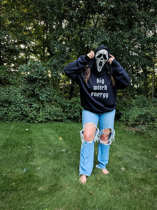 A girl wearing ripped jeans, a bedazzled ghost face mask, and a black sweatshirt that says "big witch energy" in a white gothic font