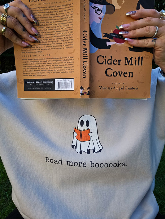 A girl covering her face with a book, wearing a tan Halloween crewneck sweatshirt featuring a ghost reading a book, with the phrase "Read more boooooks" underneath