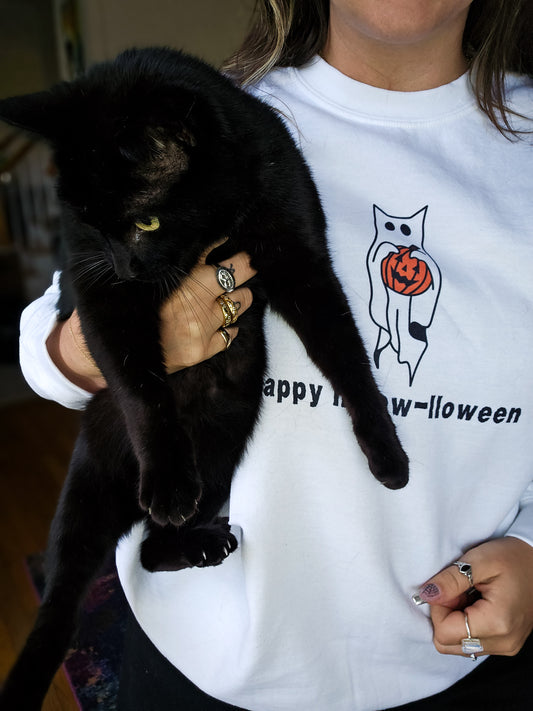 A girl wearing a white Halloween crewneck sweatshirt featuring a ghost with cat ears, holding a jack-o-lantern, with the phrase "Happy Meow-lloween" underneath. The girl is also holding a real black cat.