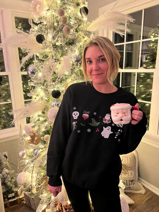 A girl standing in front of a Christmas tree, wearing a black crewneck sweater with an array of spooky-inspired Christmas images