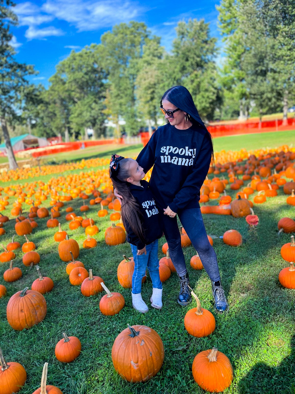 A mom and her daughter standing in a pumpkin patch, each wearing a black sweatshirt. One sweatshirt says "spooky mama" and the other says "spooky babe"