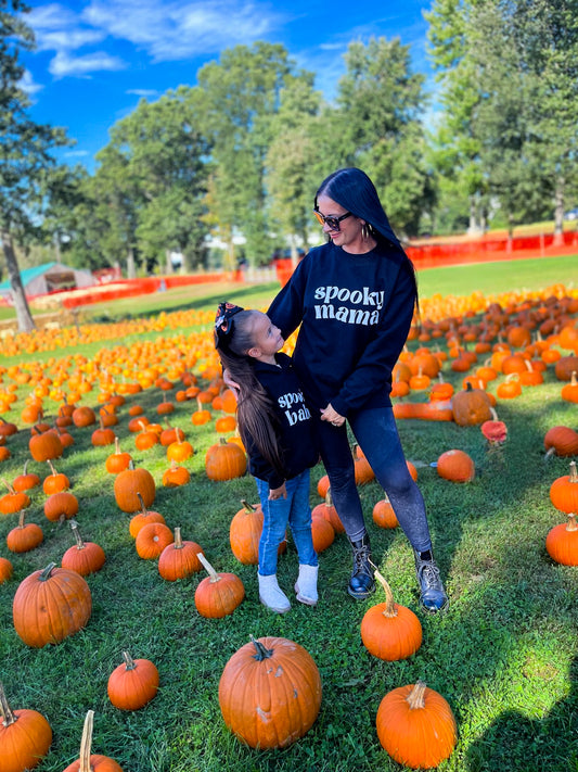 A mom and her daughter standing in a pumpkin patch, each wearing a black sweatshirt. One sweatshirt says "spooky mama" and the other says "spooky babe"