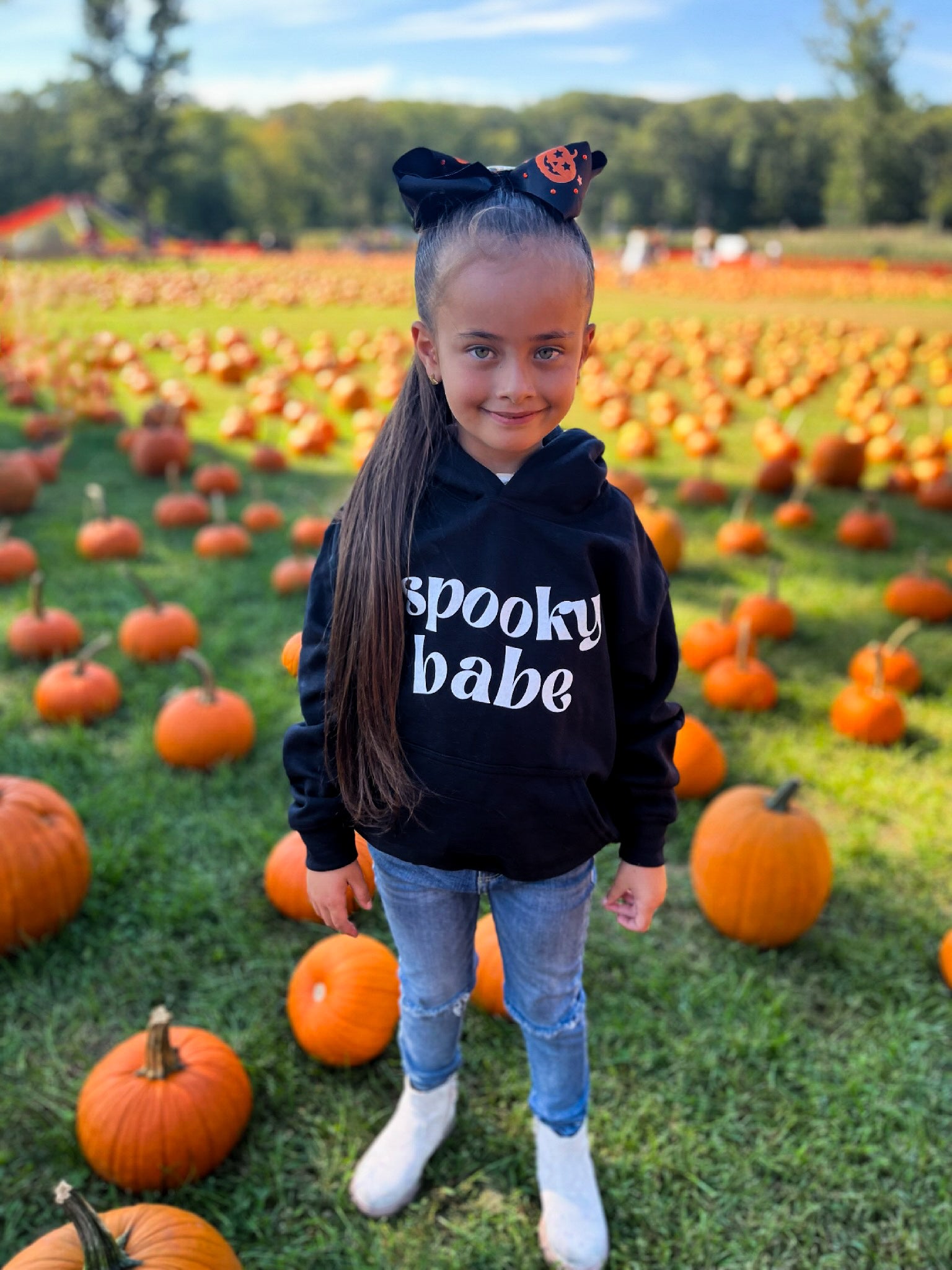 A little girl standing in a pumpkin patch, wearing a black hoodie that says "spooky babe" in a trendy white font