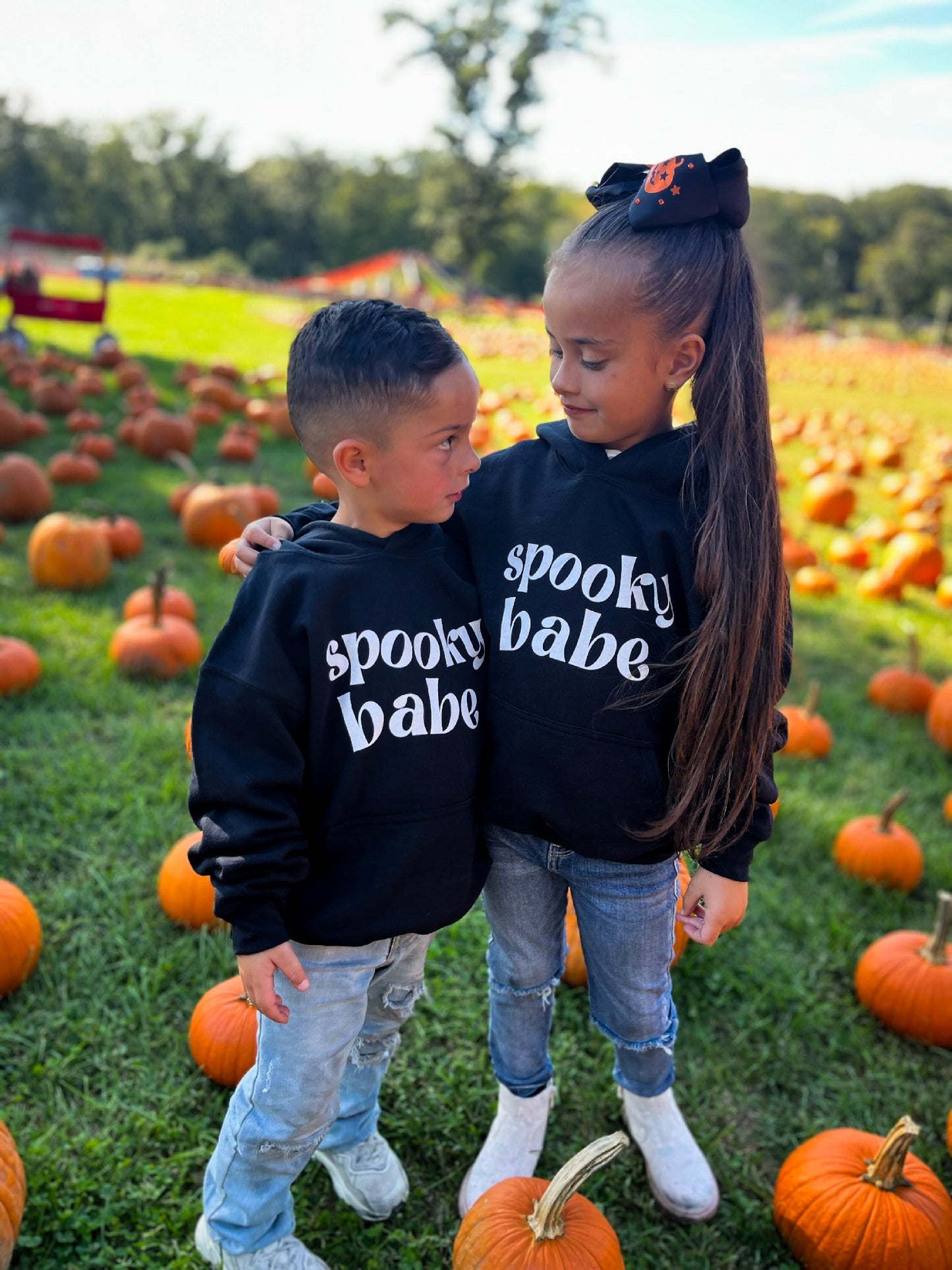 A little boy and girl standing in a pumpkin patch, each wearing a black hoodie that says "spooky babe" in a trendy white font