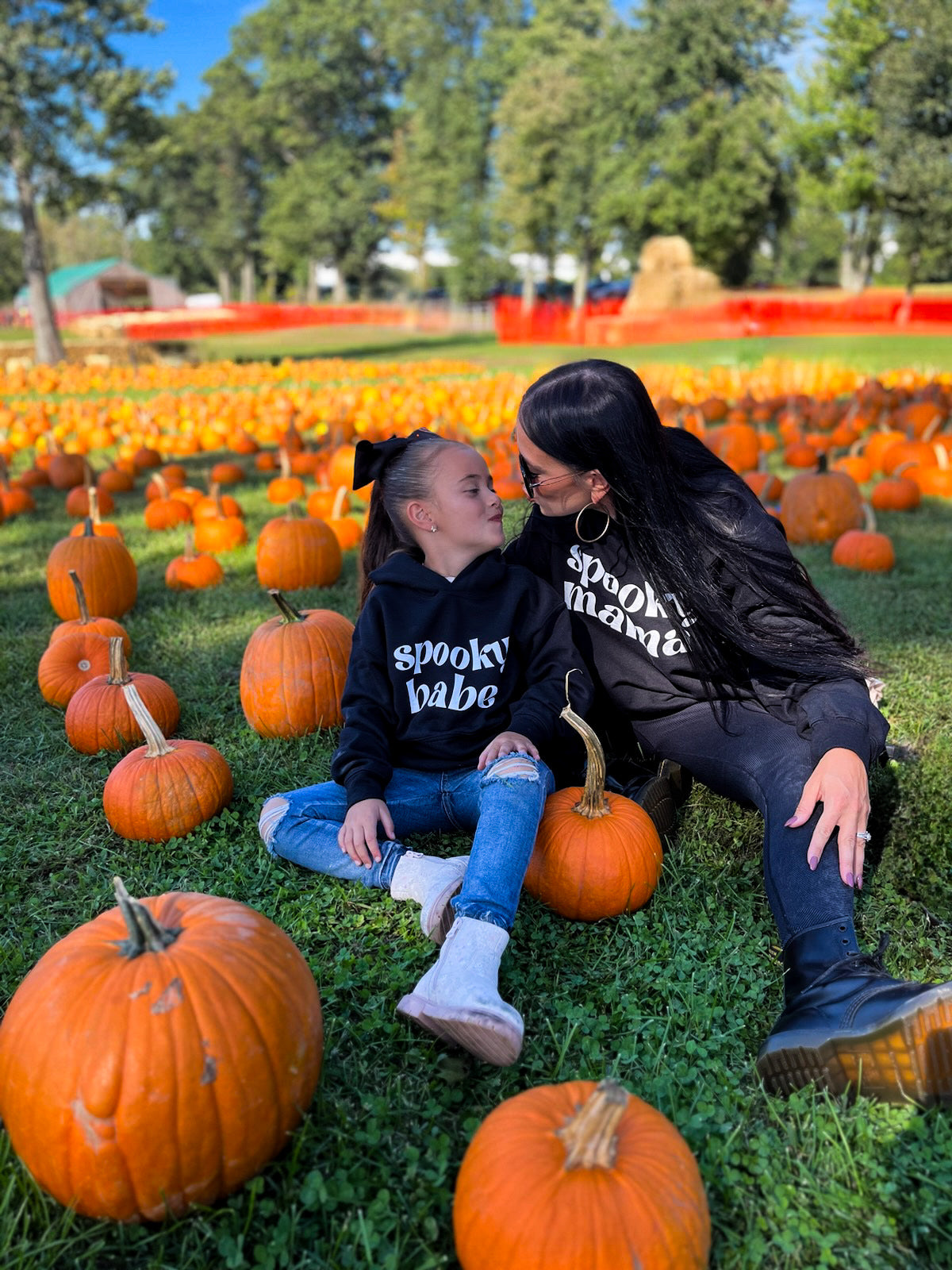 A mom and her daughter sitting in a pumpkin patch making kissy faces at each other, wearing matching black sweatshirts that say "spooky mama" and "spooky babe"