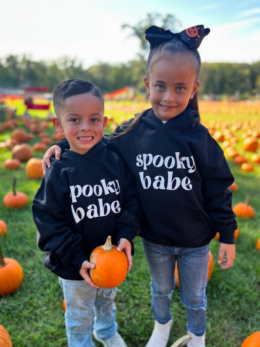 A little boy and girl standing in a pumpkin patch, each wearing black hoodies that say "spooky babe" in a trendy white font