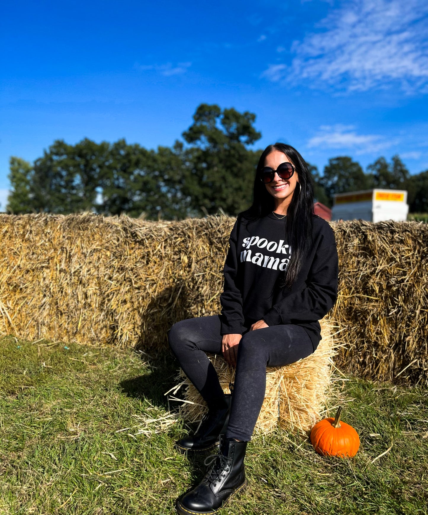 A girl sitting on a hay bale, wearing black leggings, black combat boots, and a black crewneck sweatshirt that says "spooky mama" in a trendy white font