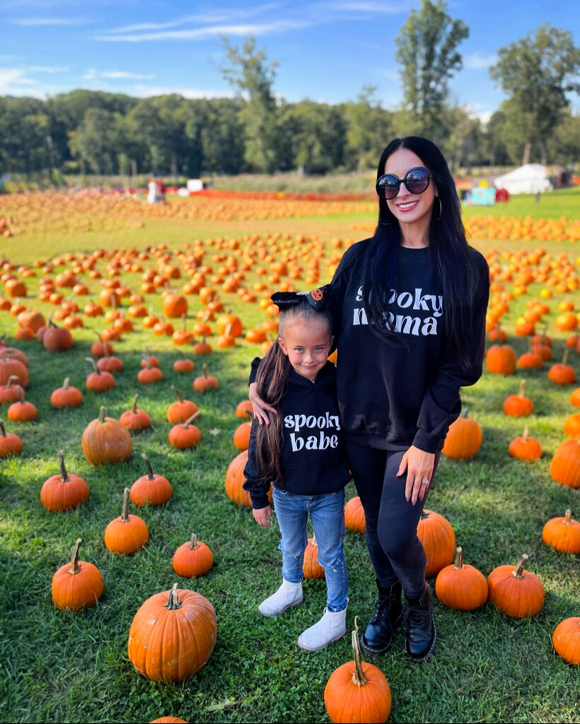 A mom and daughter standing in a pumpkin patch, each wearing matching black sweatshirts, one that says "spooky mama" and the other "spooky babe" in a trendy white font