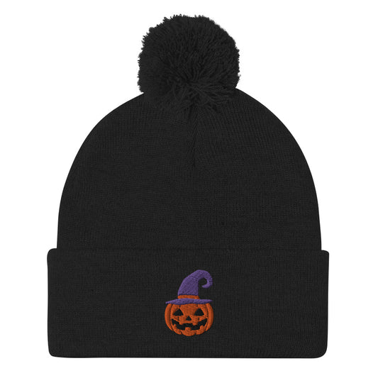 A black cuffed Halloween beanie with a pom-pom on top, featuring an embroidered jack-o-lantern wearing a purple witch hat