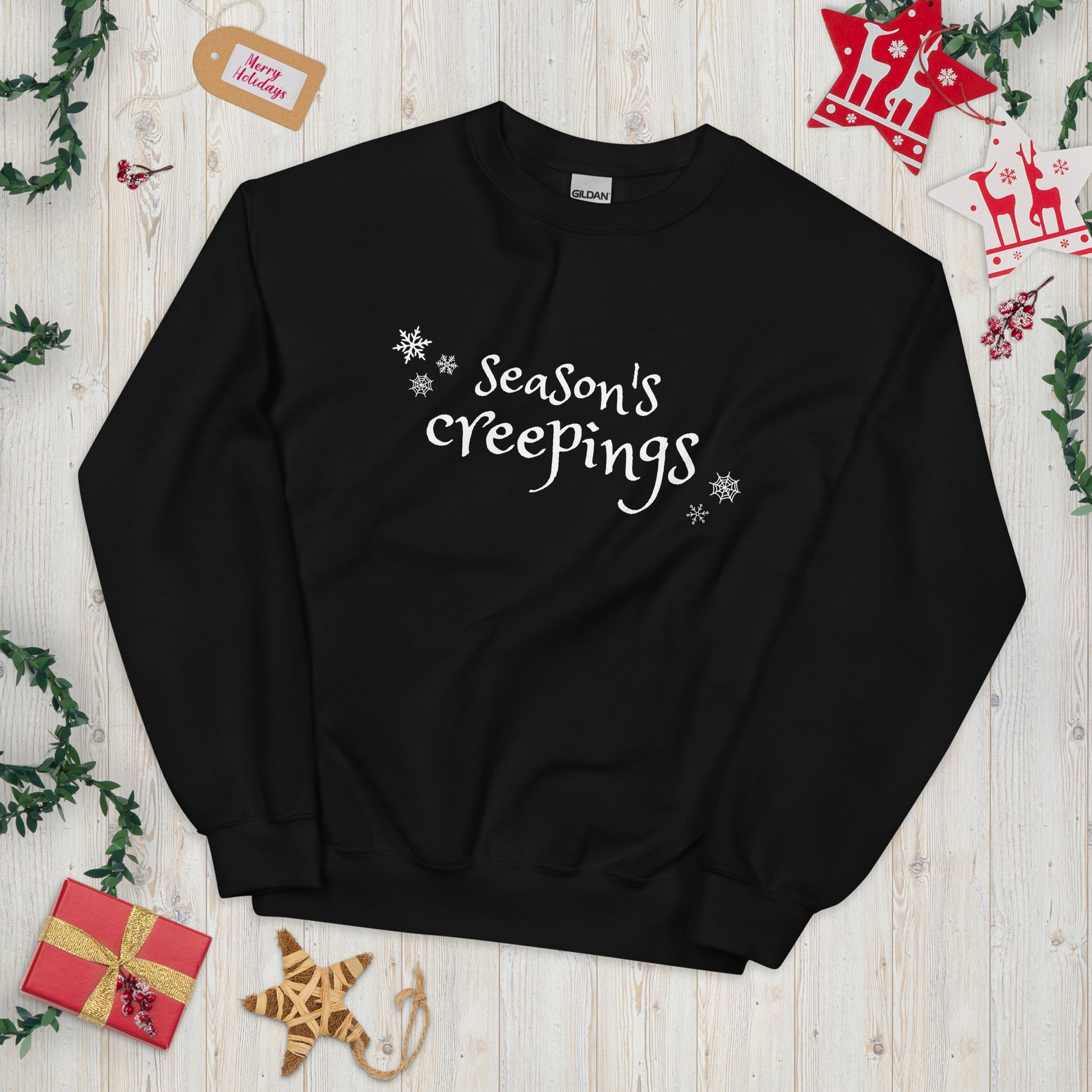 A black crewneck sweatshirt with the phrase Season's Creepings written in a whimsical white font, surrounded by snowflakes and spider webs