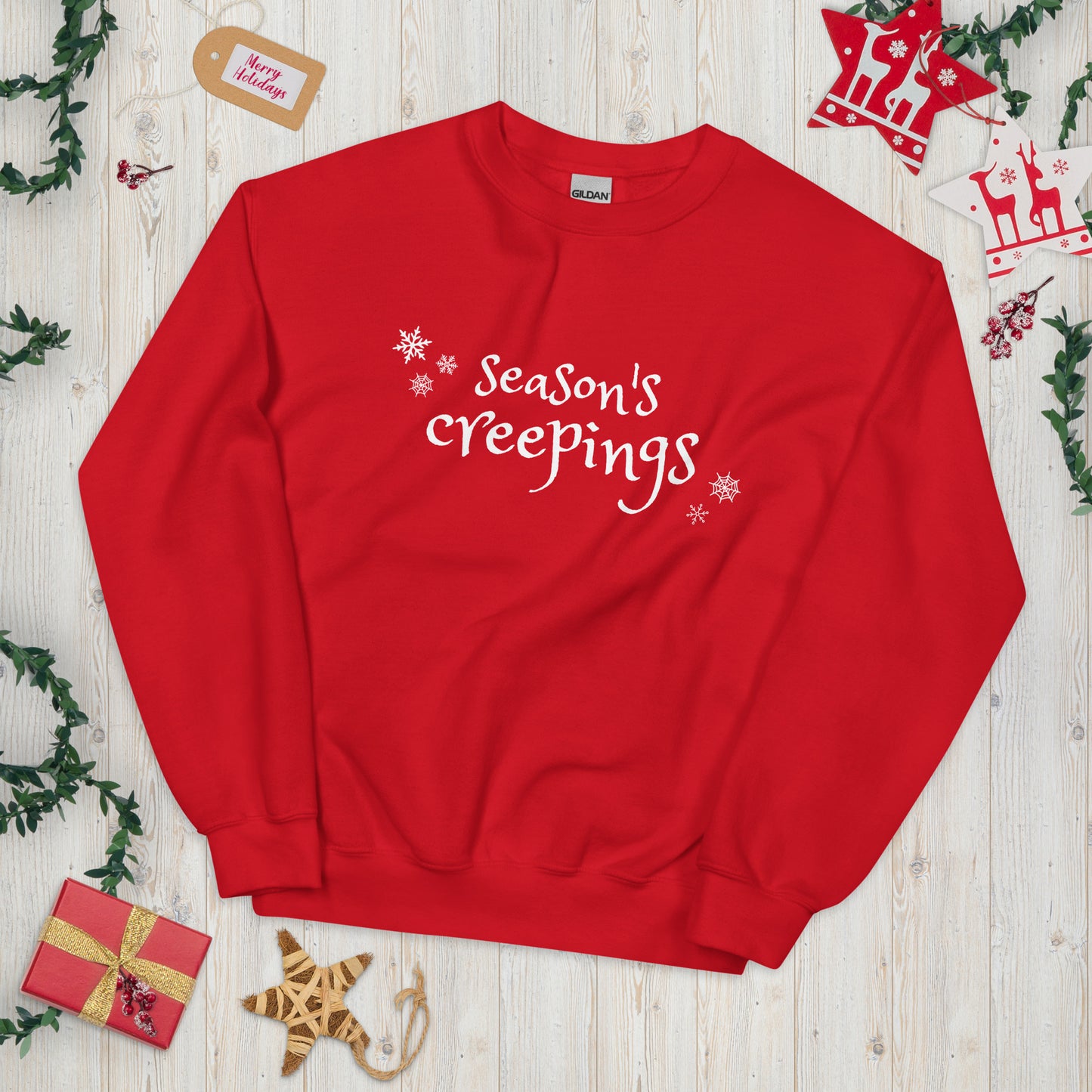 A red crewneck sweatshirt with the phrase Season's Creepings written in a whimsical white font, surrounded by snowflakes and spider webs