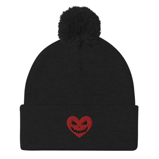 A black pom-pom beanie featuring an embroidered red heart with a jack-o-lantern face