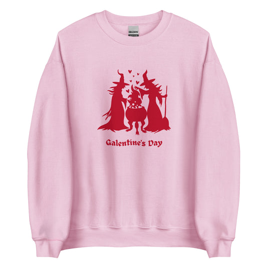 A pink crewneck sweatshirt featuring a red silhouette of two witches standing over a cauldron with heart bubbles coming out. The phrase Galentine's Day is written in red gothic letters underneath the silhouette.